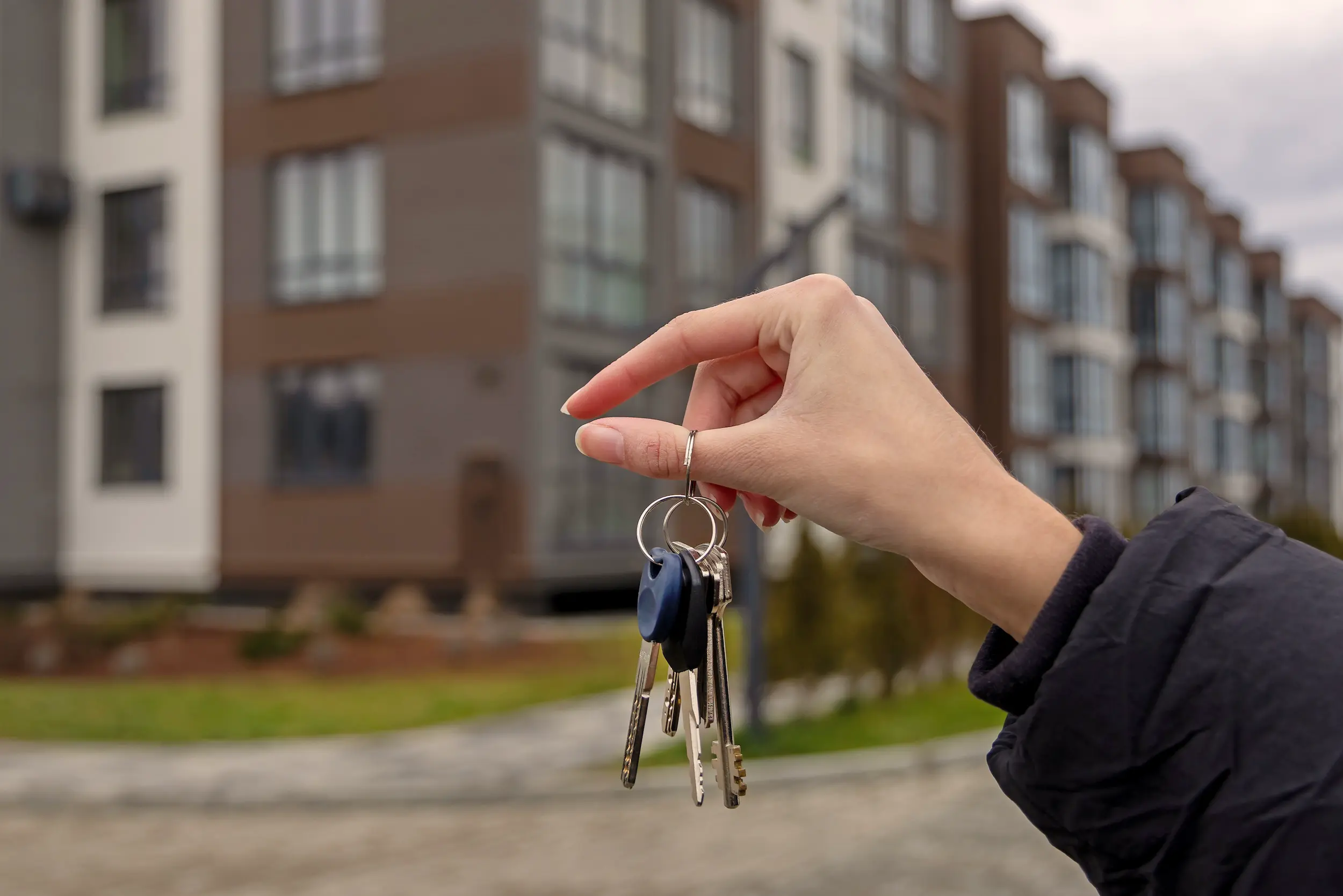 Wohnungsschlüssel werden vor einem Wohnkomplex durch eine Hand hochgehalten.
